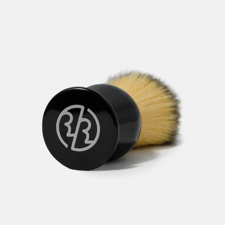 The Rockwell Synthetic Shaving Brush by Rockwell Razors features soft beige bristles, a black handle with a white logo, and is ideal to pair with your classic razor. Photographed against a plain light background.