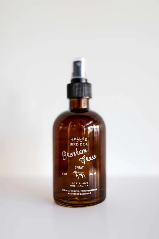 A brown glass 8 oz bottle labeled "Room & Linen Spray | Brenham Grass | Ballad of the Bird Dog" features a black spray nozzle. Made with essential oils, it stands out against a white background and is perfect for freshening rooms or linens.