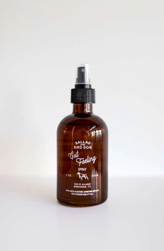 A brown glass spray bottle labeled "Room & Linen Spray | Gut Feeling | Ballad of the Bird Dog" by Ballad of the Bird Dog, with a black plastic pump, filled with essential oils, stands on a white surface against a plain background.