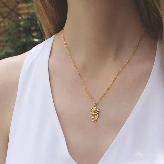 A person wears a white sleeveless top and the Rose with Stem Necklace by Vintage Acorn, featuring a 16K gold-plated rose pendant and a nickel- and lead-free gold-filled clasp.