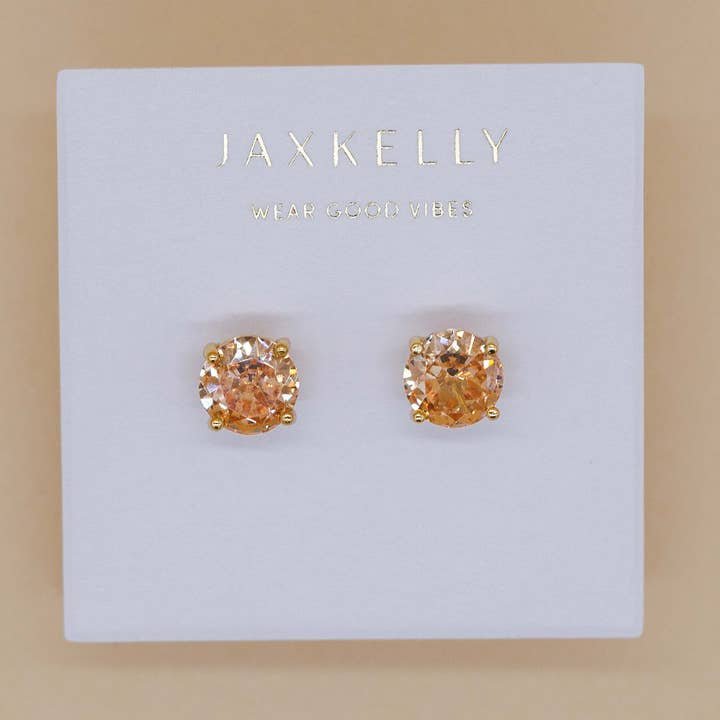 A pair of Round Cz Champagne stud earrings by JaxKelly with orange stones and 18k gold-plated settings, displayed on a white JaxKelly card.