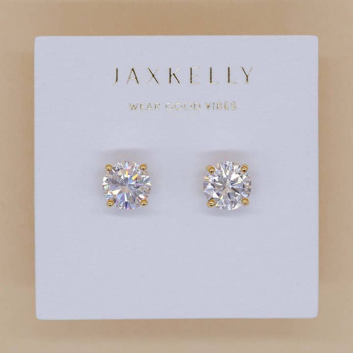 The JaxKelly Round CZ Earrings feature 18k gold-plated studs on a white card with "WEAR GOOD VIBES." Hypoallergenic for comfortable, everyday wear.
