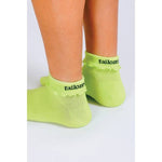 A person stands on a light surface wearing Tailored Union’s Ruffle Ankle Sock in light green with reinforced heel and "tailored" in black lettering near the cuff.