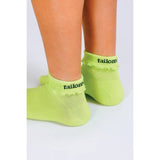 A person stands on a light surface wearing Tailored Union’s Ruffle Ankle Sock in light green with reinforced heel and "tailored" in black lettering near the cuff.