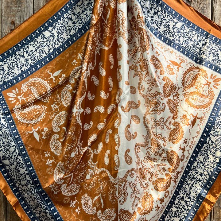 The Rodeo Drive Rust & Navy Paisley Wild Rag features floral designs in brown, white, and navy blue. Displayed on a wooden surface, this western scarf adds elegant style to any outfit.