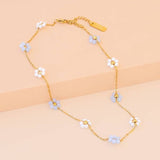 The Sabrina Necklace by Peter and June features 18K gold plating with small blue and white flower charms accented by sparkling zircon stones, elegantly arranged along the chain and displayed on a beige surface.