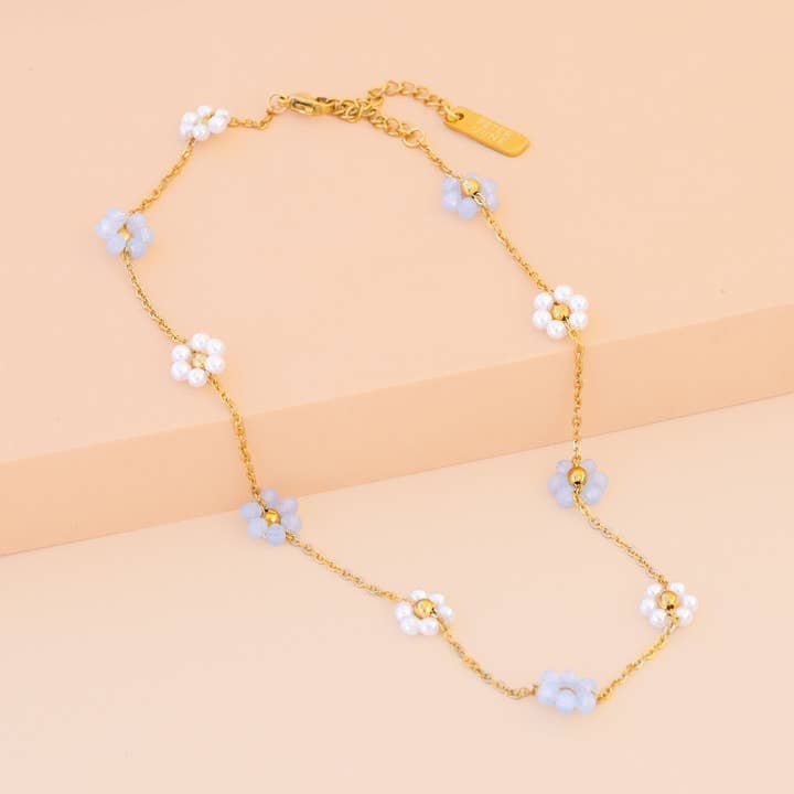 The Sabrina Necklace by Peter and June features 18K gold plating with small blue and white flower charms accented by sparkling zircon stones, elegantly arranged along the chain and displayed on a beige surface.