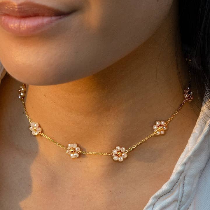A close-up of a person wearing the Sabrina Necklace by Peter and June, featuring 18k gold plating, small pearl flower charms, and sparkling zircon stones, paired with a light-colored top.
