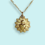 The Sacred Heart Necklace by Ornamental Things features a radiant sunburst and raised sacred heart in a vintage-inspired gold-plated design, shown here against a light blue background.