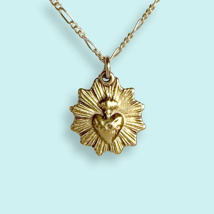 The Sacred Heart Necklace by Ornamental Things features a radiant sunburst and raised sacred heart in a vintage-inspired gold-plated design, shown here against a light blue background.