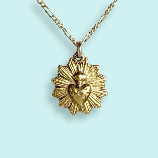 The Sacred Heart Necklace by Ornamental Things features a gold-plated, vintage-inspired sacred heart with a flame detail, handmade in the USA and shown against a light blue background.