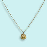 The Sacred Heart Necklace by Ornamental Things features a gold-plated sunburst design with a heart at the center, vintage-inspired pendant, and is pictured on a light blue background.