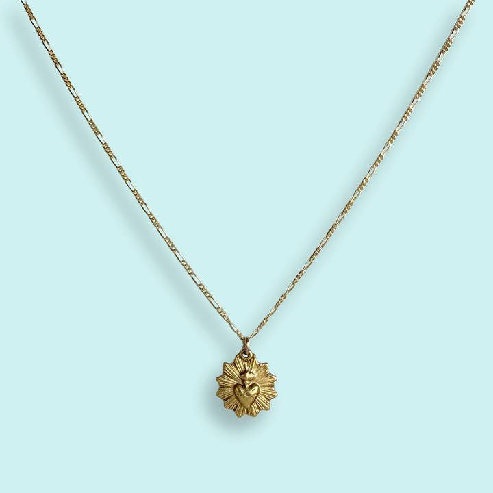 The Sacred Heart Necklace by Ornamental Things features a gold-plated sunburst design with a heart at the center, vintage-inspired pendant, and is pictured on a light blue background.