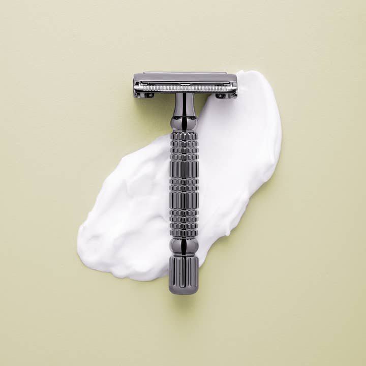The Rockwell Razors Safety Razor | R1 sits on a layer of white shaving cream against a light beige background, offering a smooth, eco-friendly shave.