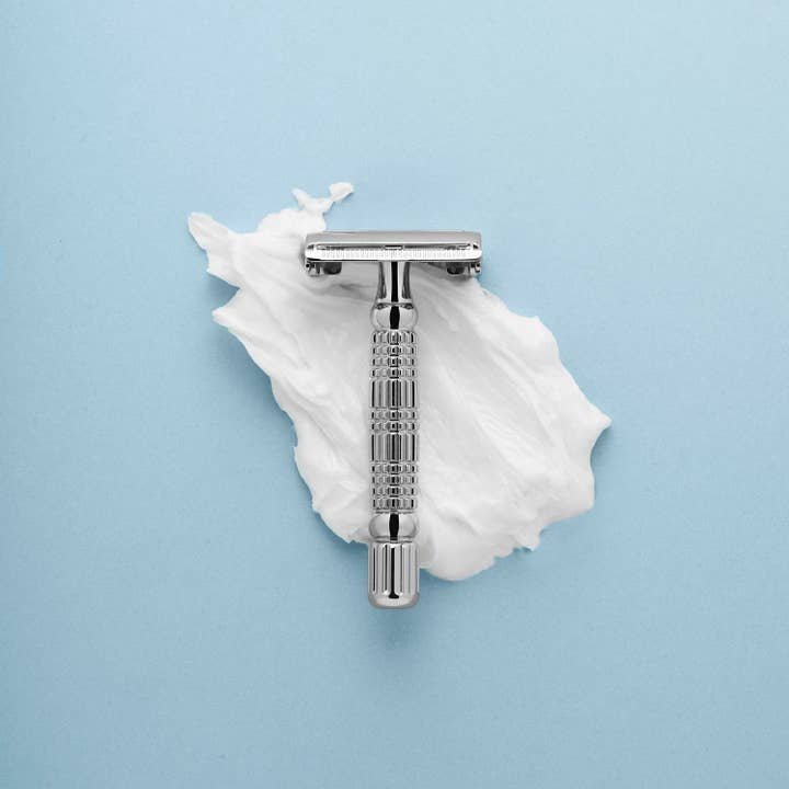 The Rockwell Razors Safety Razor | R1, a silver single blade razor, rests on white shaving cream against a light blue background, offering a smooth shave and an eco-friendly plastic-free experience.