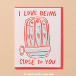 The Sardine Love Card by And Here We Are features sardines in a tin with "I love being close to you" in red and orange neon ink—a quirky love card for someone special.
