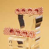 Two Satsuma Orange Gingham Acetate Retro Circle Alligator Clips by Winona Irene, featuring orange and white checkered cellulose acetate with circular indents, are displayed on clear acrylic blocks against a yellow background.