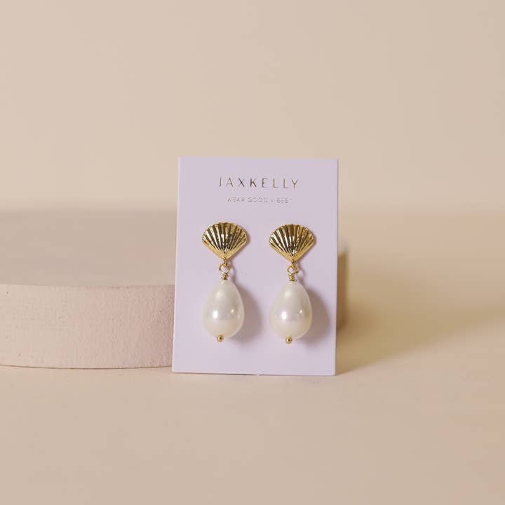 Seashell Pearl Drop Gold Earrings by JaxKelly, hypoallergenic and tarnish-free, feature gold seashells with dangling white pearls. Displayed on a branded card against a beige background.