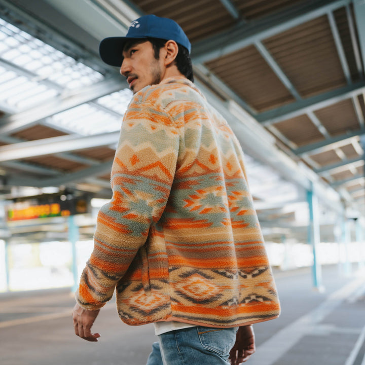 A man in the Sendero Provisions Co. Sedona Fleece, blue cap, and jeans walks through a bright urban space with metal beams.