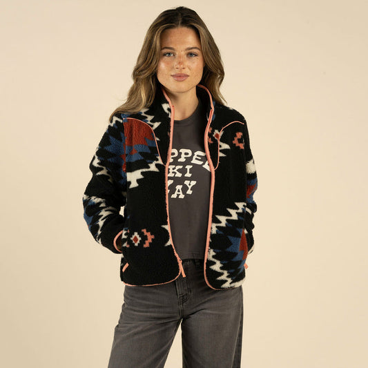 A woman with long hair wears the Sedona Fleece by Sendero Provisions Co. over a dark graphic t-shirt, standing with her hands in her pockets against a plain beige background.
