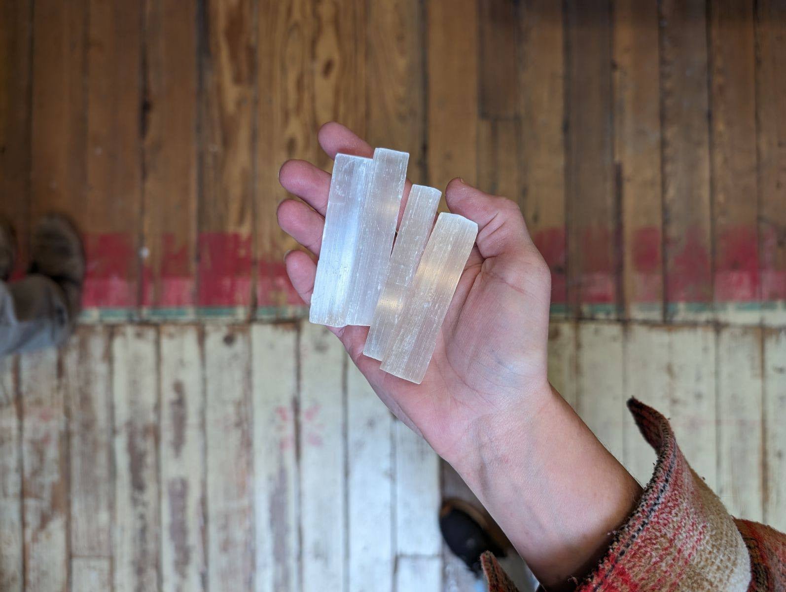 A hand holds three translucent Selenite Crystal Stacks by World Incense Store above a worn wooden floor with a red strip, perfect for chakra work and crystal healing.