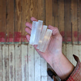 A hand holds three translucent Selenite Crystal Stacks by World Incense Store above a worn wooden floor with a red strip, perfect for chakra work and crystal healing.