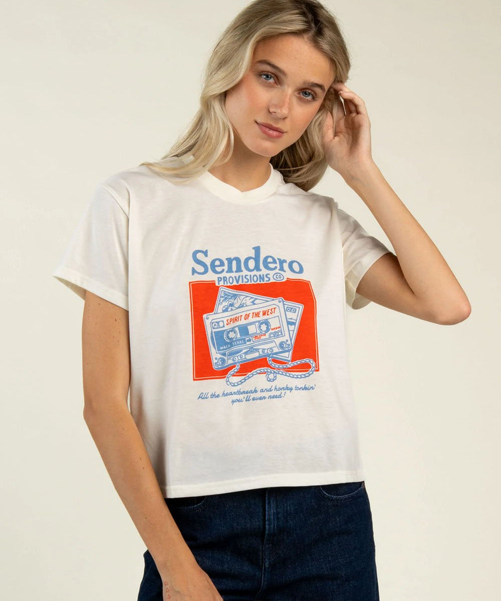 A woman wearing the Cassette Tape Boxy Tee from Sendero Provisions Co.—a white, ultra-soft, modestly cropped graphic T-shirt—paired with dark jeans stands against a plain background, touching her hair for an effortlessly stylish look.