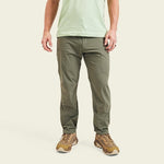 A person wearing olive green Shoalwater Tech Pants by Howler Brothers, a light green t-shirt, and brown athletic shoes stands against a plain background.
