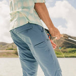 Person in a light plaid shirt and blue Shoalwater Tech Pants by Howler Brothers is fly fishing near water, holding a fishing rod with mountains and sky visible in the background.