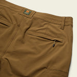 Close-up of the back of tan Shoalwater Tech Pants by Howler Brothers, showing two back pockets (one zippered) and a visible Howler Brothers label above the pockets.