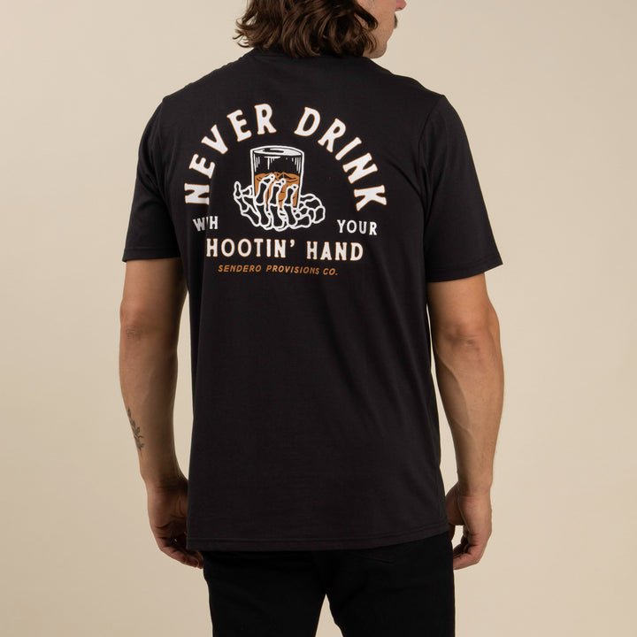 A person wears Sendero Provisions Co.'s Shootin' Hand black ultra-soft cotton tee, featuring a soft touch screen print of a skeleton hand with a drink and "Never Drink With Your Hootin' Hand" on the back.