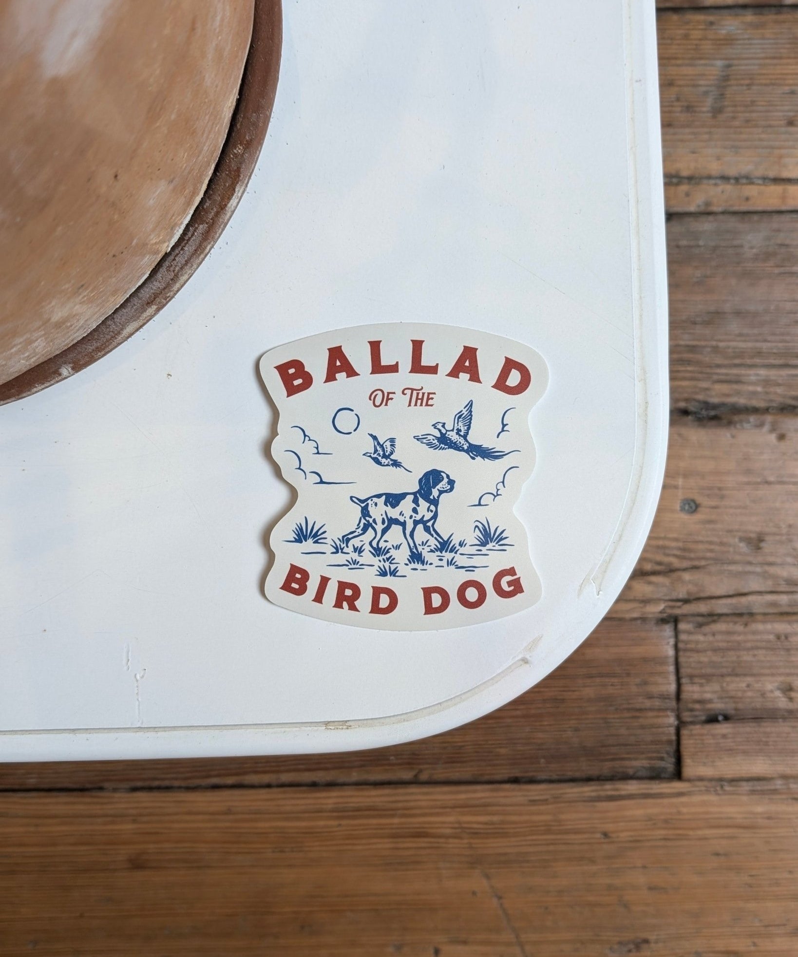 The Ballad of the Bird Dog Shop Cooler Sticker | Pheasant Flush features a pointing dog, birds, plants, and clouds in blue and red. Durable for outdoor use, it stands out on any surface—perfect for coolers or adventures.