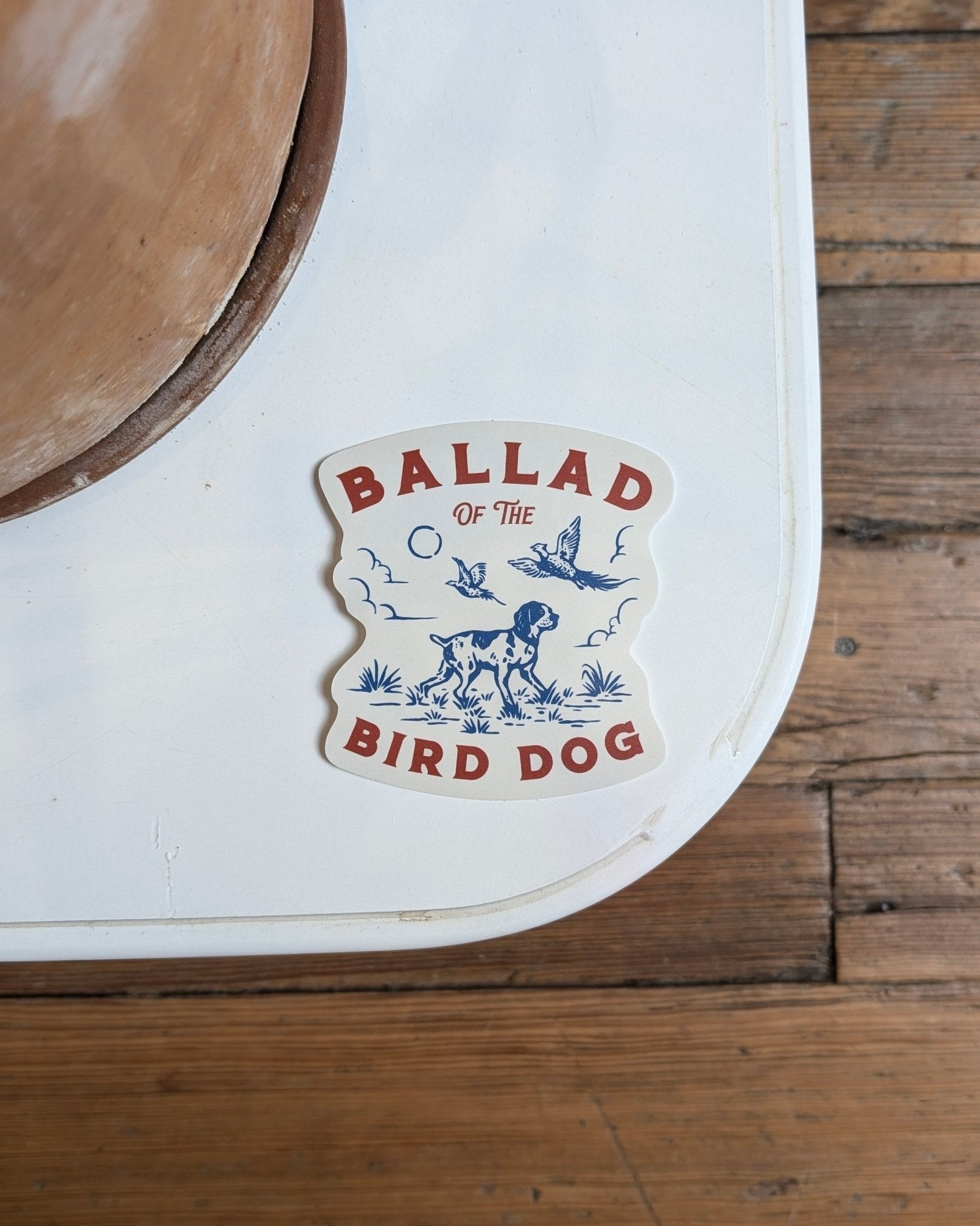 The Ballad of the Bird Dog Shop Cooler Sticker | Pheasant Flush features a pointing dog, birds, plants, and clouds in blue and red. Durable for outdoor use, it stands out on any surface—perfect for coolers or adventures.