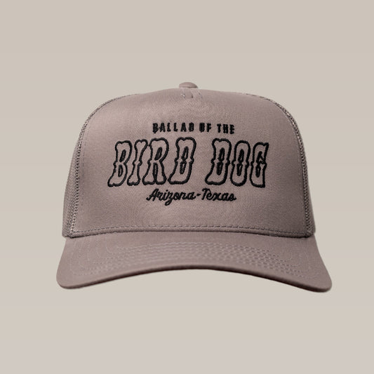 The Ballad of the Bird Dog Shop Hat features gray fabric with black “Arizona-Texas” text on the front, shown against a beige background.