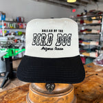 The Ballad of the Bird Dog Shop Hat | Arizona to Texas is a white and black cap displayed indoors on a wooden stand, featuring the "Ballad of the Bird Dog" brand name.