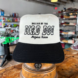 The Ballad of the Bird Dog Shop Hat | Arizona to Texas is a white and black cap displayed indoors on a wooden stand, featuring the "Ballad of the Bird Dog" brand name.