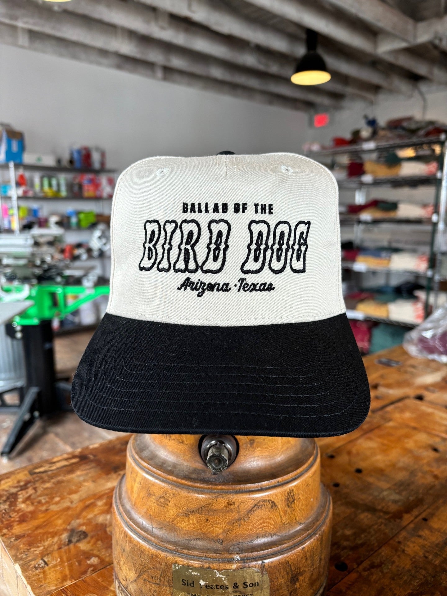 The Ballad of the Bird Dog Shop Hat | Arizona to Texas is a white and black cap displayed indoors on a wooden stand, featuring the "Ballad of the Bird Dog" brand name.
