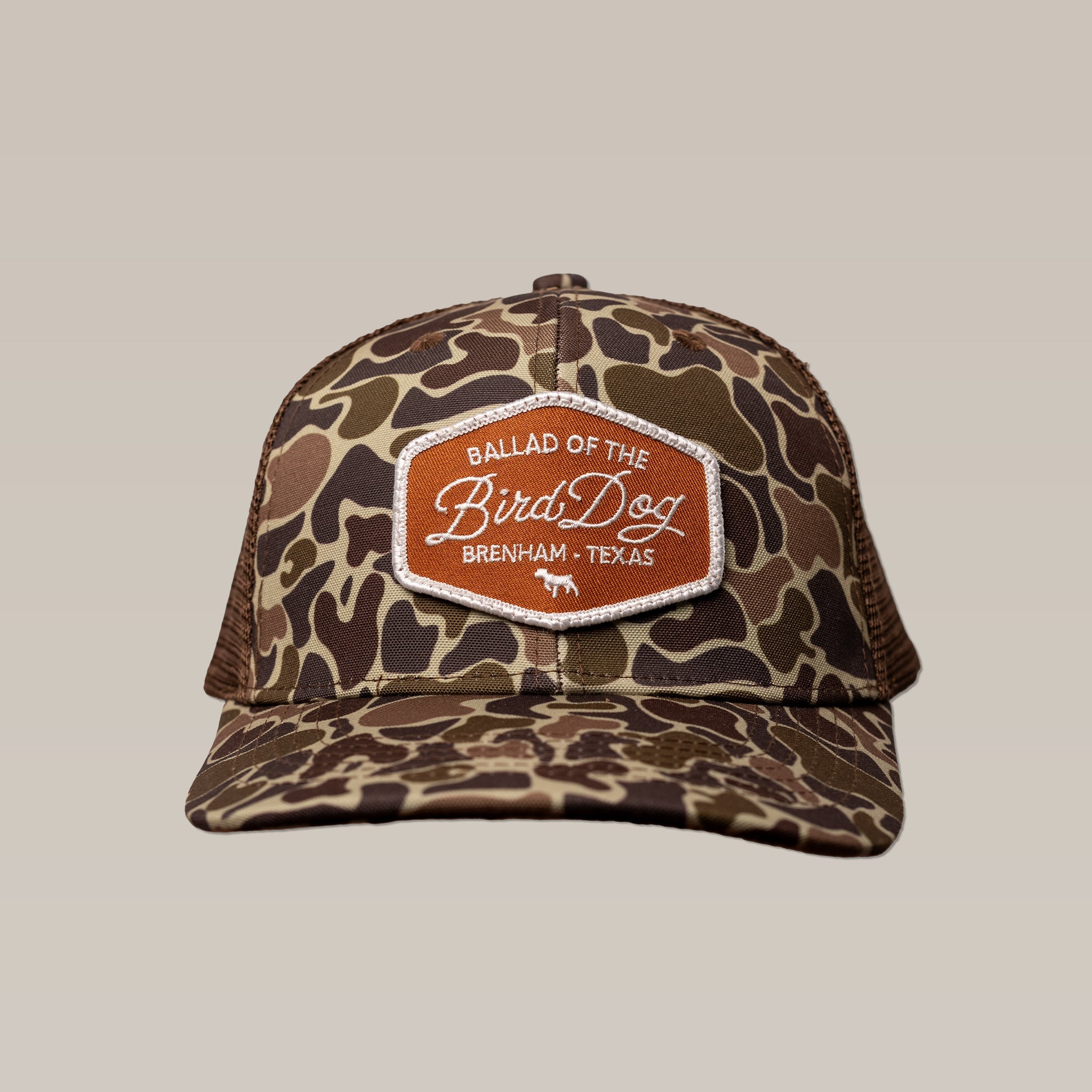 The Ballad of the Bird Dog Shop Hat | Badge features a camouflage design with an orange "Ballad of the Bird Dog, Brenham, Texas" patch, shown against a plain backdrop.