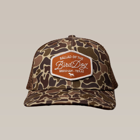The Shop Hat | Badge by Ballad of the Bird Dog is a camo trucker hat featuring an orange patch with "Ballad of the Bird Dog, Brenham, Texas" on the front.