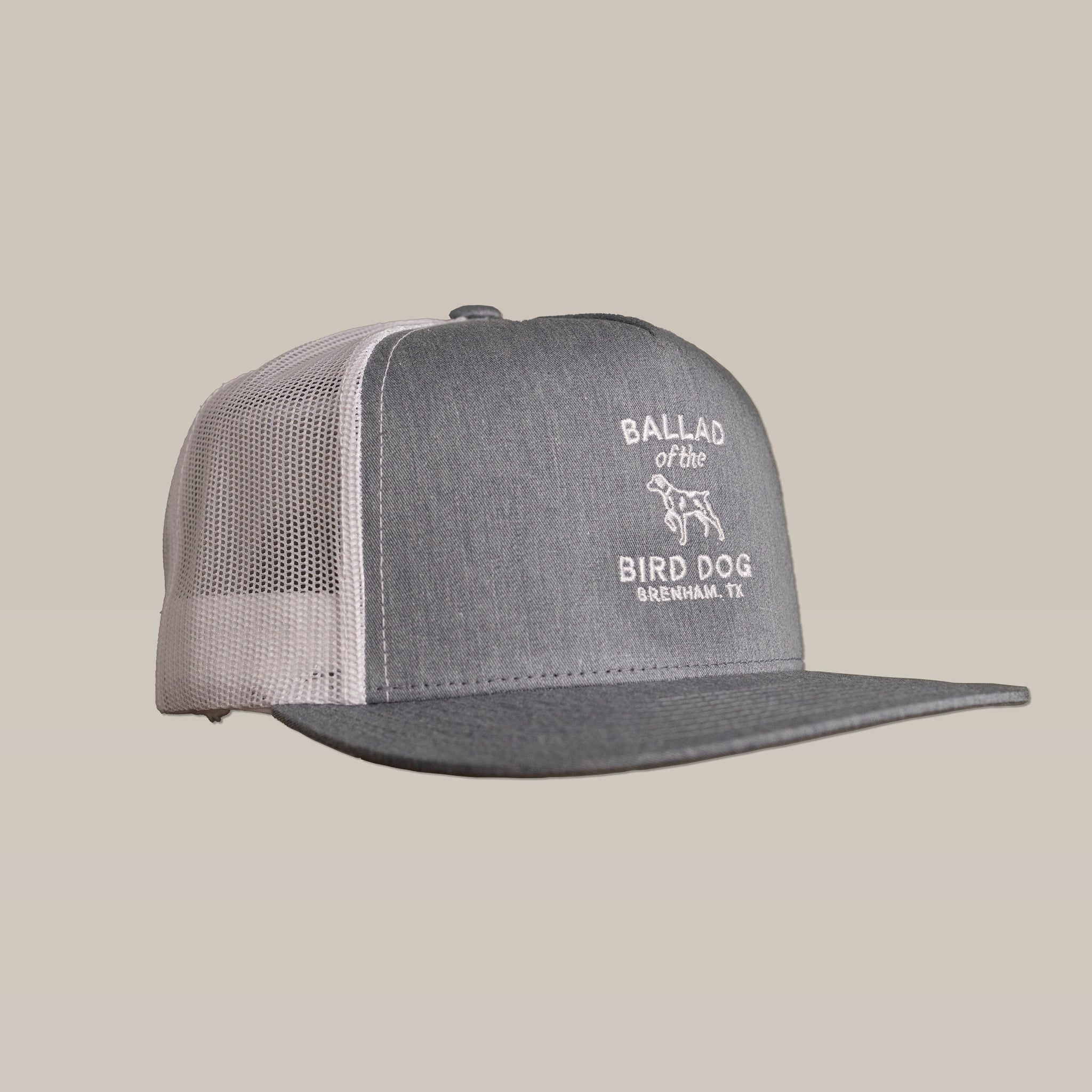 The Shop Hat | Bird Dog Classic Shop by Ballad of the Bird Dog is a gray and white mesh trucker hat with a dog graphic and "Ballad of the Bird Dog Brenham, TX" logo embroidered on the front—a classic for quality goods fans.