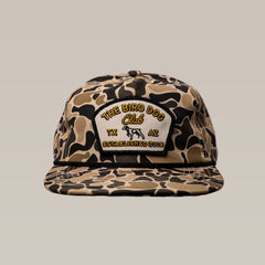 A camouflage baseball cap with a front patch reading “THE BIRD DOG Club TX AZ ESTABLISHED 2018” and featuring an illustrated dog.