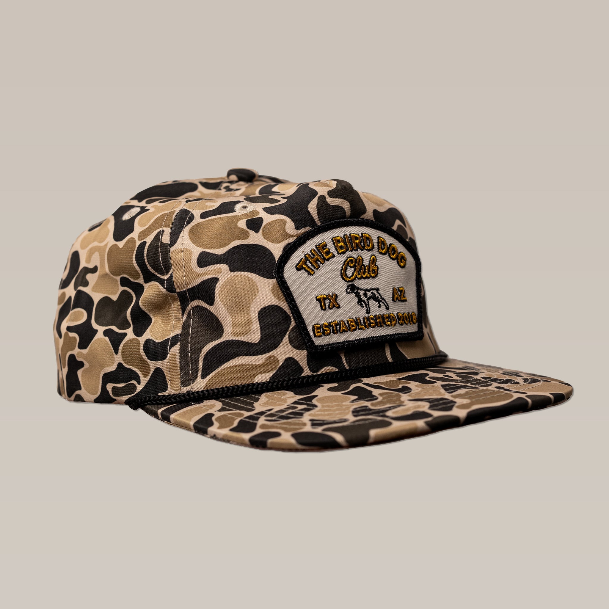 The Shop Hat | Bird Dog Club by Ballad of the Bird Dog is a camo snapback with a front patch reading "The Bird Dog Club TX AZ Established 1984"—heritage apparel for outdoor enthusiasts. Pictured on a plain beige background.