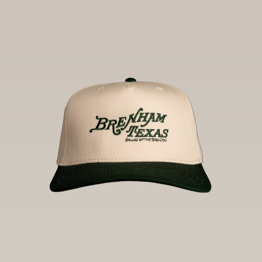 The Shop Hat by Ballad of the Bird Dog features "Brenham Texas Heritage Script" and "Ballad of the Bird Dog" embroidery in green on a beige and dark green cap—ideal for fans of classic style and local pride.