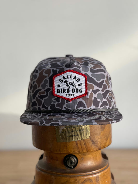 The Shop Hat | Camo Companion by Ballad of the Bird Dog features a gray camo print and a red and white "Ballad of the Bird Dog Texas" patch, perfect as an outdoor hat, displayed on a wooden stand.