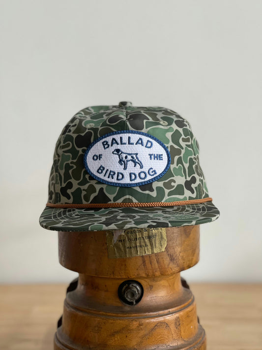 The Ballad of the Bird Dog Shop Hat | Camo Emblem is displayed on a wooden hat stand.