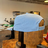 The Ballad of the Bird Dog Shop hat, featuring the Classic Shop Logo in light blue, is displayed on a wooden hat form in a workshop with equipment visible in the background.
