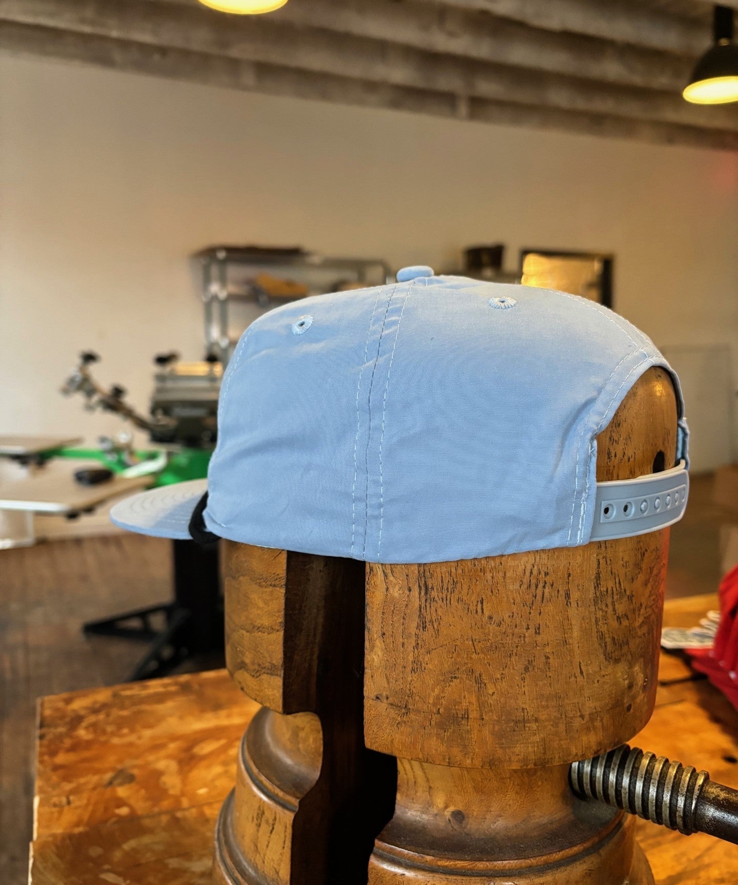 The Ballad of the Bird Dog Shop hat, featuring the Classic Shop Logo in light blue, is displayed on a wooden hat form in a workshop with equipment visible in the background.