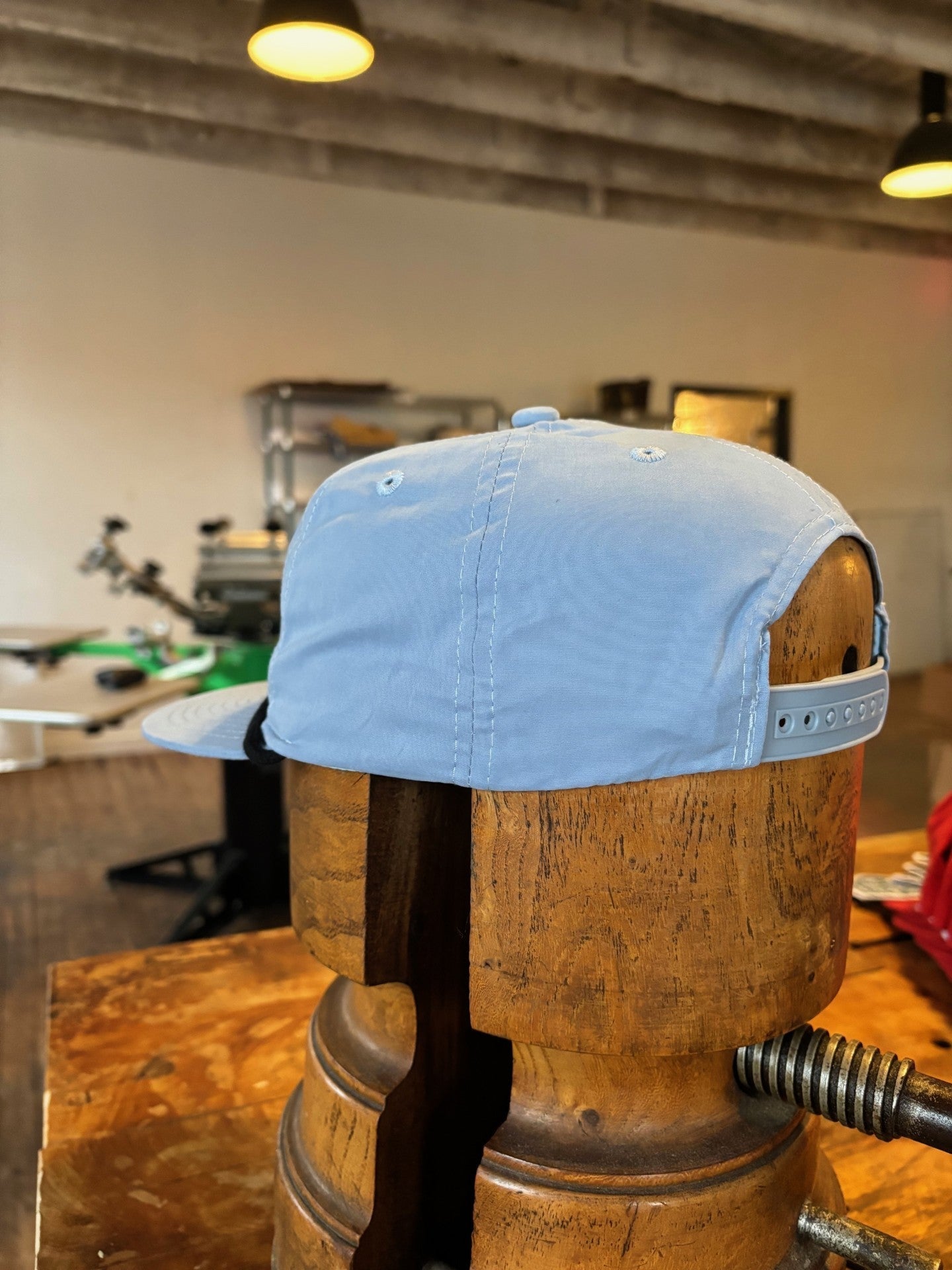 The Ballad of the Bird Dog Shop hat, featuring the Classic Shop Logo in light blue, is displayed on a wooden hat form in a workshop with equipment visible in the background.