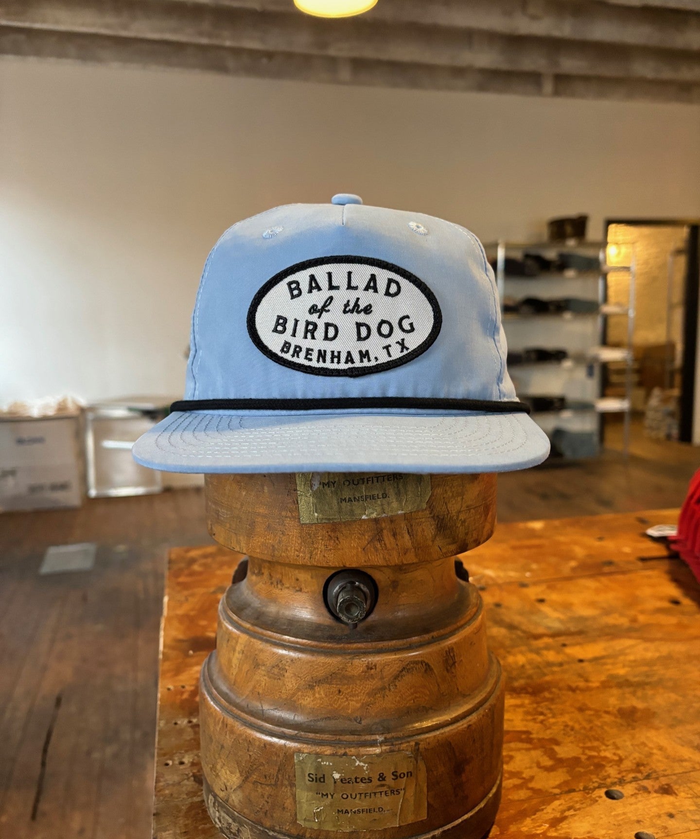 The Shop hat by Ballad of the Bird Dog, featuring the Classic Shop Logo patch, is showcased on a vintage wooden hat block in a rustic shop.