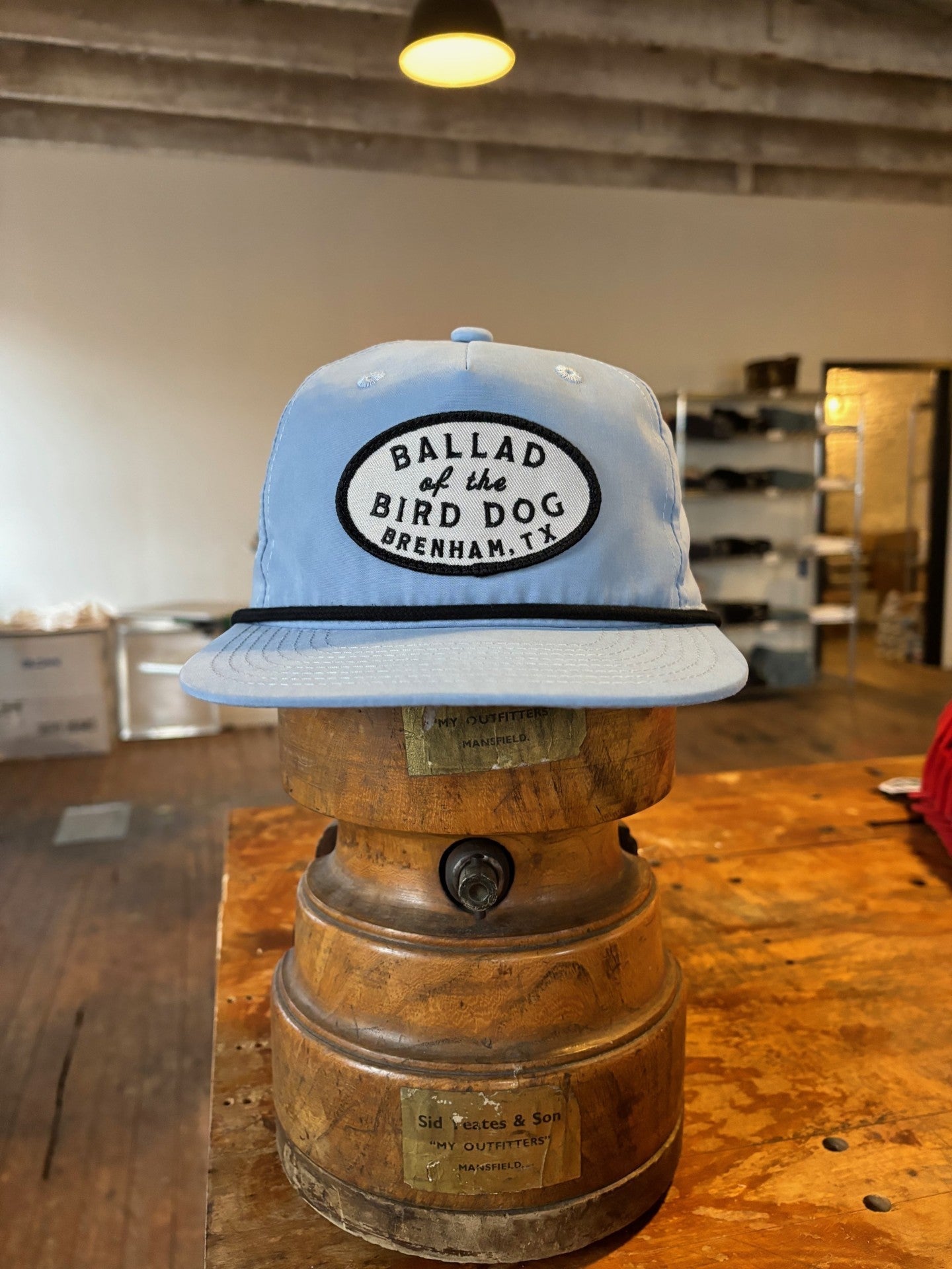 The Shop hat by Ballad of the Bird Dog, featuring the Classic Shop Logo patch, is showcased on a vintage wooden hat block in a rustic shop.